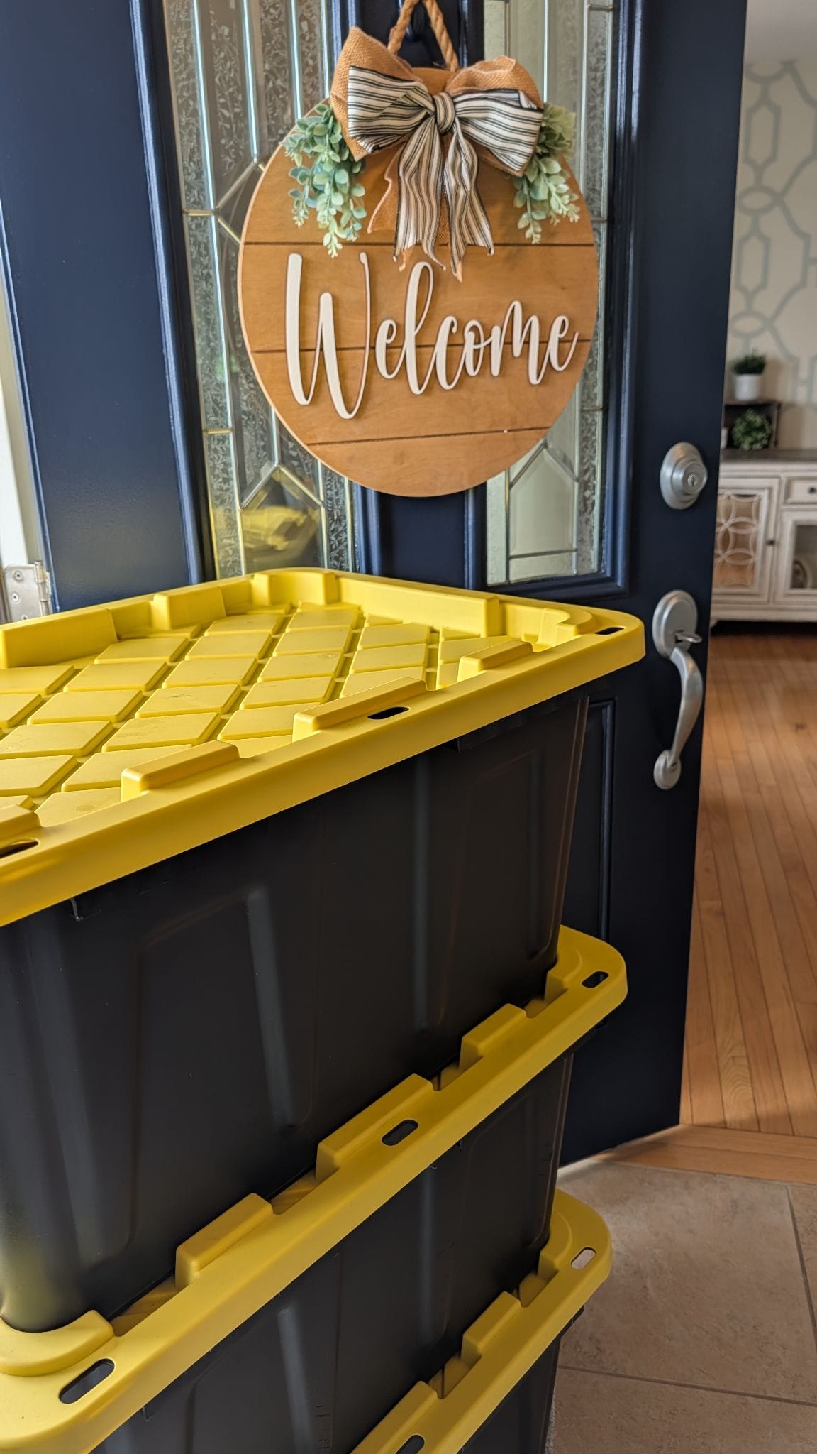 Two black Crate & Key moving totes with bright yellow lids stacked just inside an open navy front door, below a wooden Welcome wreath.