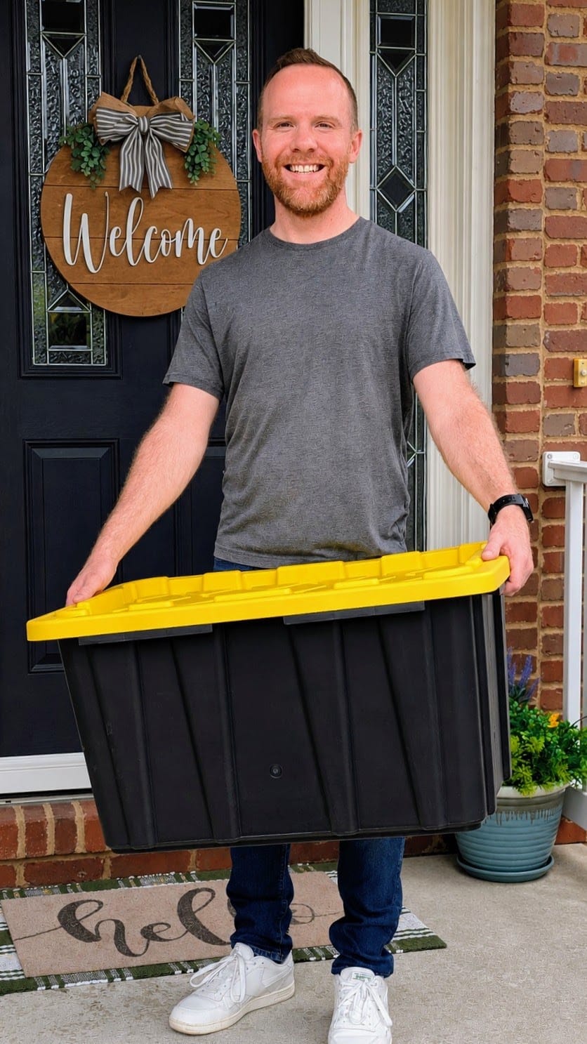 Ryan, the owner of Crate & Key, smiling on a front porch while holding a black moving tote with a yellow lid.