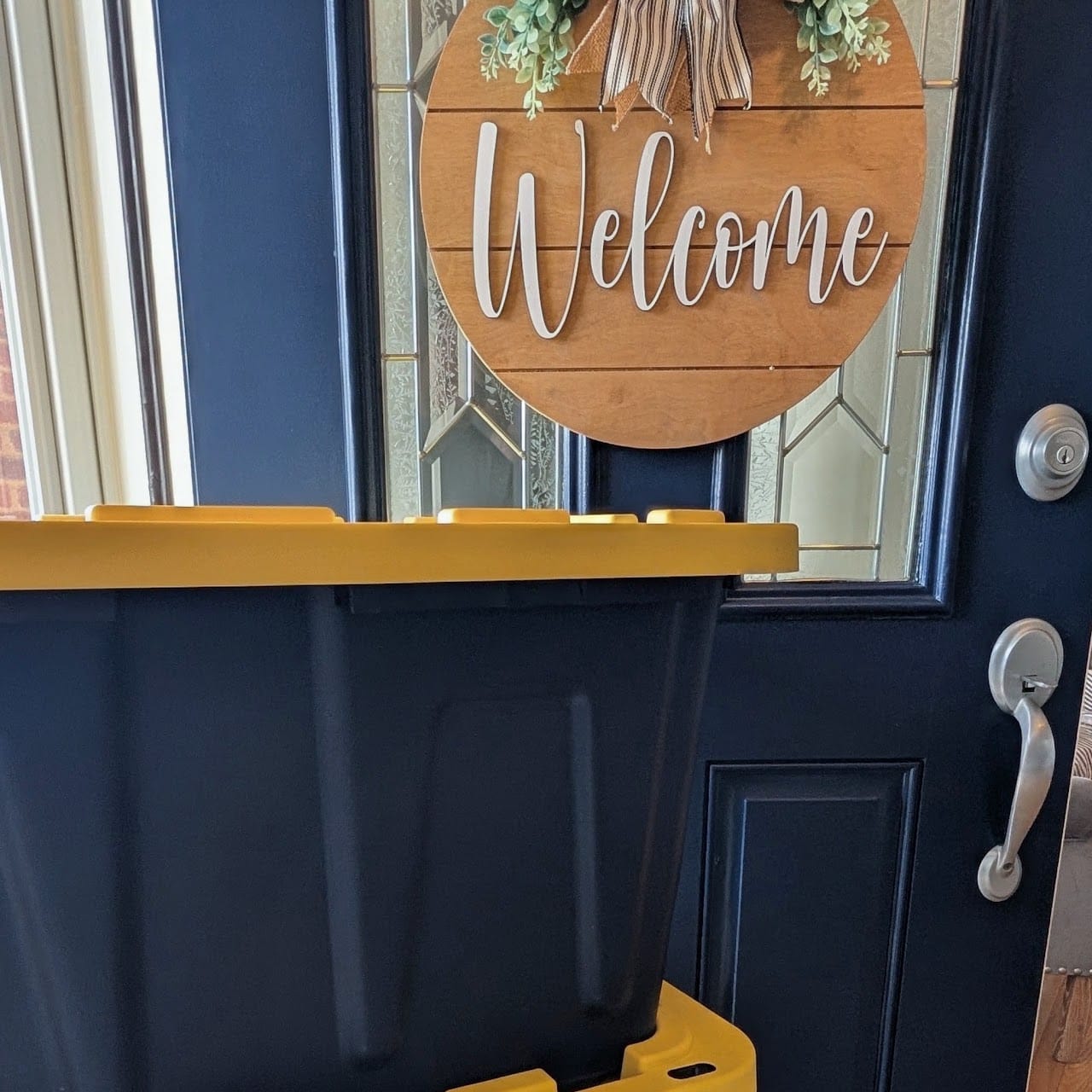 A black tote with a yellow lid waiting at a navy front door beneath a wooden Welcome wreath, ready for collection.