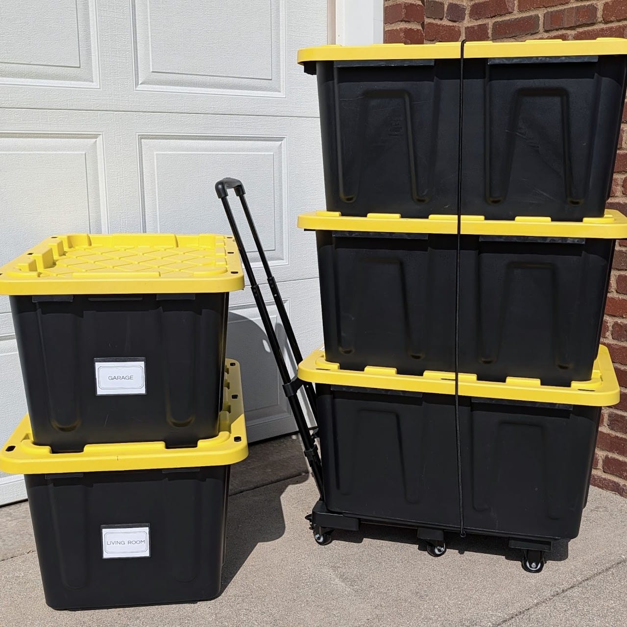 Stacked black totes with yellow lids on a hand truck by a garage door, labelled GARAGE and LIVING ROOM.